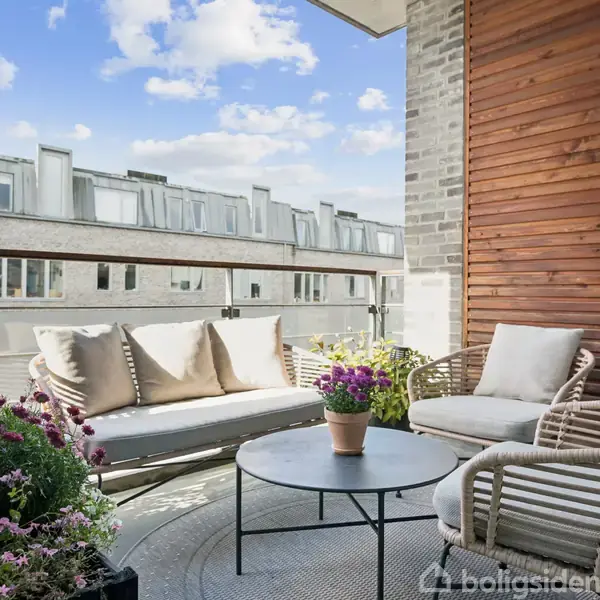 Sofa og stole står på en altan, omgivet af blomster i krukker. Solsken oplyser området, og en murstensvæg med trælister giver en hyggelig atmosfære. Udsigt til nabobygninger.