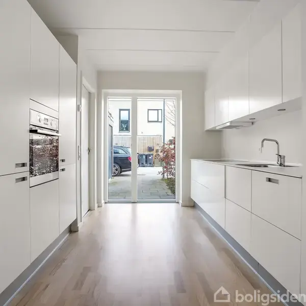 Et moderne køkken med hvide kabinetter og indbygget ovn. En vask er placeret på højre side. Gennemsigtigt dørparti åbner ud til en gårdhave med lidt grønt og en bil udenfor.