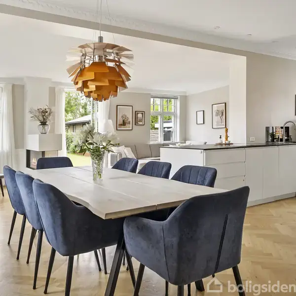 Et lyst spisebord med blå polstrede stole står i en elegant spisestue. En moderne lampe hænger over bordet. Rummet har trægulv, store vinduer og et åbent køkken i baggrunden.