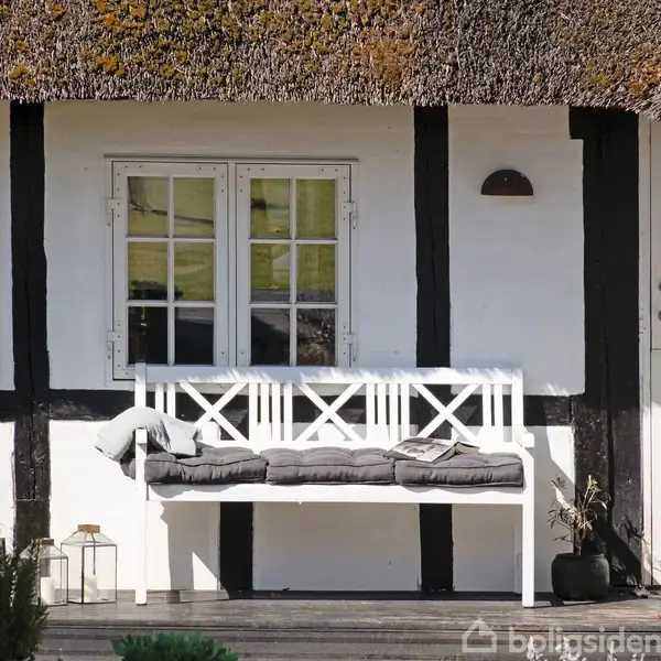 Bænk med hynder står stille foran bindingsværkshus med stråtag. Området er roligt med planter omkring, og huset har hvide vinduer og dør, hvilket skaber en idyllisk stemning.