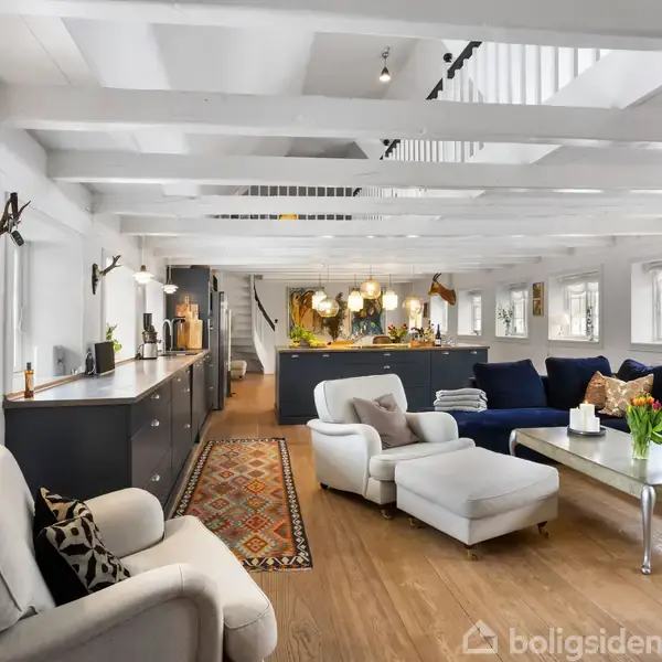 Lyse møbler og en sofa står i en moderne, åben stue med trægulve og hvide loftssprosser. Rummet har mange vinduer og indbygget belysning, med en trappe i baggrunden.