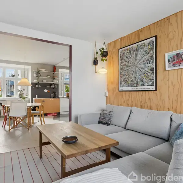 Grå sofa står i stuen, dekoreret med billeder på trævæg; et sofabord står foran. I baggrunden ses køkkenområde med spisebord og vinduer med planter.