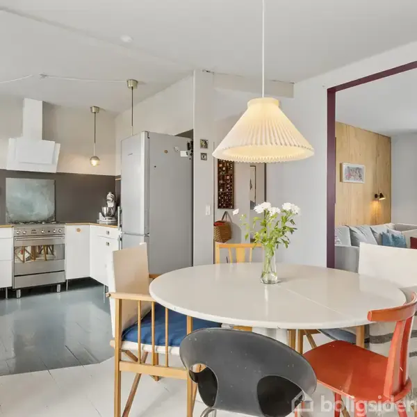 Rundt spisebord med farverige stole står i en åben stue-køkken indretning. I baggrunden ses et moderne køkken med hvide skabe og stålkomfur. Der er en sofa ved et vindue.