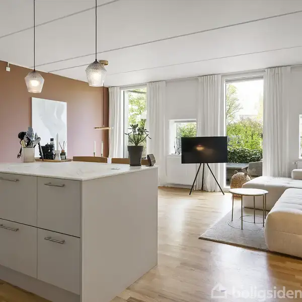 Et moderne køkken-alrum har en hvid køkkenø med opbevaring. To lamper hænger over øen. Sofa og tv er placeret i et lyst, rummeligt område med trægulv og store vinduer.