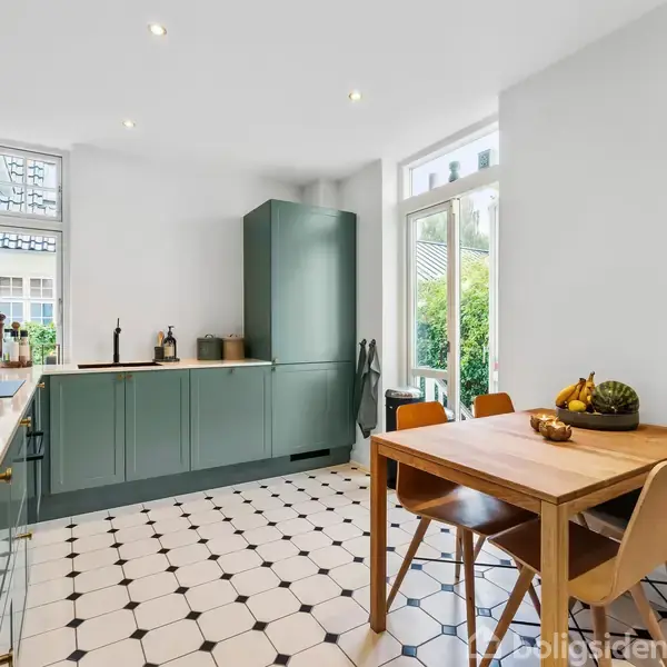 Køkken med grønne skabe og en træbord med frugtskål. Aktion er ikke til stede, og konteksten inkluderer hvide vægge, sorte og hvide fliser samt store vinduer med udsigt til have.