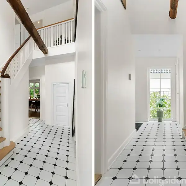 Trappe med trætrin fører opad i en lys, hvidmalet gang med sorte, mønstrede fliser. Der er synlige træbjælker i loftet og en grøn plante ved et vindue bagerst.