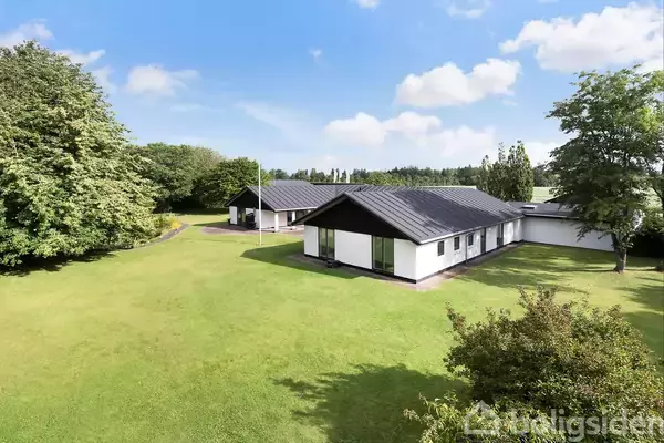 En hvid enfamilieshus med sort tag står på en stor, velholdt græsplæne omgivet af træer under en blå himmel med lette skyer.