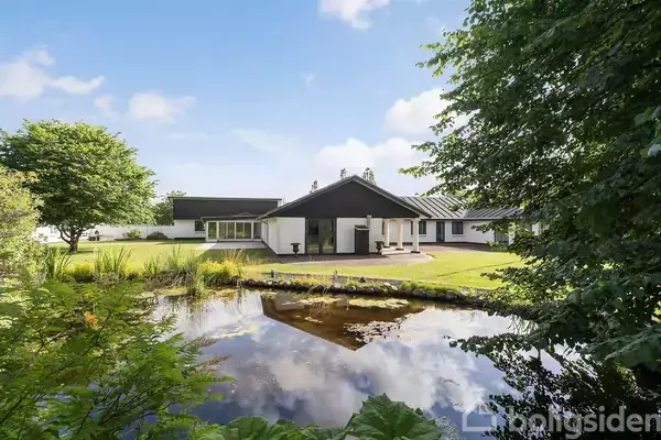 Et moderne hus står ved en rolig dam i en frodig have. Dammen spejler himlen og omkringliggende træer. Huset har hvide vægge og et sort tag.