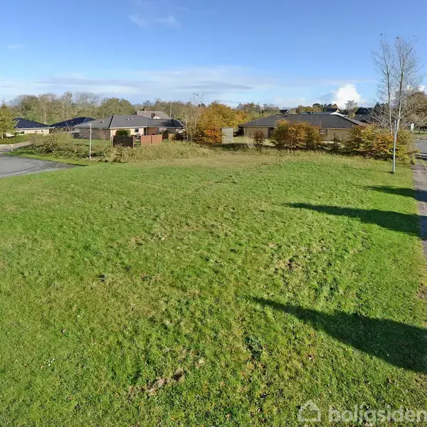 Grøn græsplæne ligger stille i et boligområde med huse og træer i baggrunden under en klar blå himmel. En vej løber til højre for plænen.