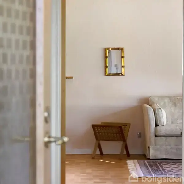 Dør åbner ind til stue med sofa i beige nuance. Et lille bord står ved siden af sofaen. En lille ramme hænger på væggen. Rummet har lyse vægge og parketgulv.