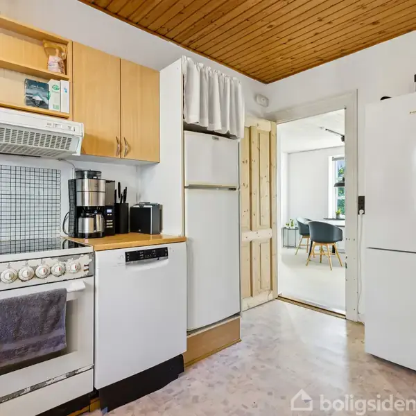 Komfur med tørklæde hængende, kaffemaskine og opvaskemaskine ved siden af, i et lyst køkken med træloft. En åben dør fører til en spisestue med stole og bord.