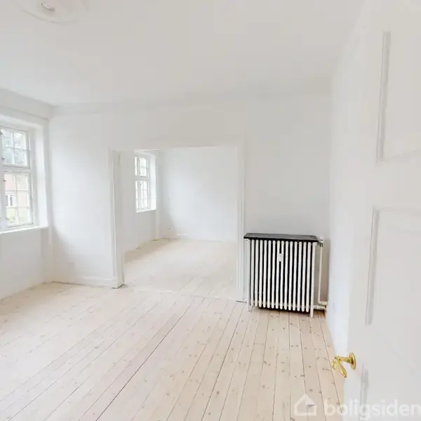 Et lyst rum med et sort radiator står ved væggen. Trægulve giver en varm atmosfære. Store vinduer tillader rigeligt med dagslys. En åbning fører til et andet rum.