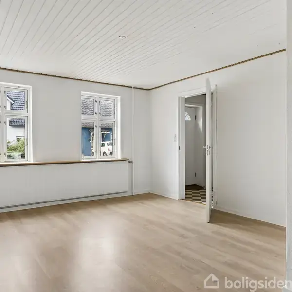 Værelse med lyse trægulve, hvide vægge og loft. To store vinduer lukker naturligt lys ind. En åben dør fører til et andet rum.