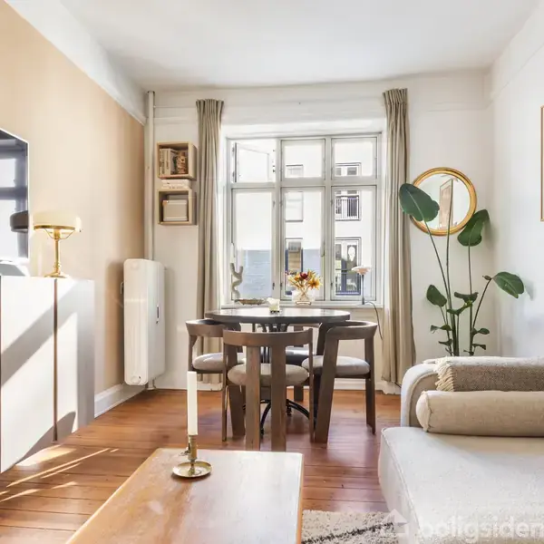 Stue med lys sofa, rund spisebord og fire stole ved vindue, fladskærms-TV på væggen, gulvlampe og grøn plante ved siden af. Dekorativt maleri hænger over sofaen.
