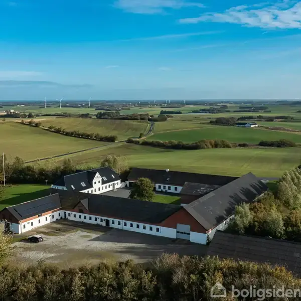 En landbrugsejendom med hvid murstensbygning og røde tage er omkranset af træer i et åbent, landligt landskab med marker strækkende sig til horisonten under en klar blå himmel.