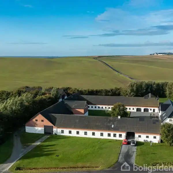 En stor gård med hvidkalkede murstensbygninger og mørkt tag omkranser en gårdsplads; placeret i et åbent, grønt landskab med marker og blå himmel i baggrunden.
