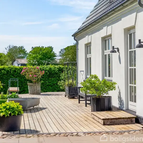 Husets terrasse er udstyret med trædæk, omkranset af grønne planter i krukker. Nabopladserne inkluderer en velholdt græsplæne og hække, mens store vinduer dominerer husets hvide facade.