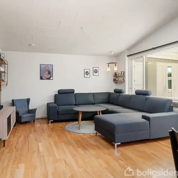 Grå hjørnesofa står i stuen med trægulv. Ved siden af er en lænestol og et TV på et lavt skab. Boghylde med dekorationer og billeder på væggen.