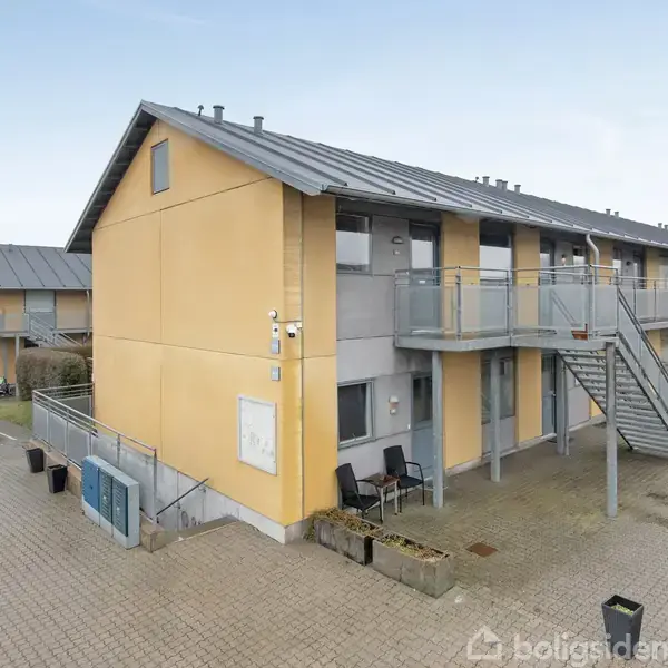 Lejlighedskompleks set udefra med to etager, flere trapper og altaner. Omkringliggende område består af flisebelagt gårdsplads med parkerede biler. Lyst, moderne arkitektur dominerer omgivelserne.