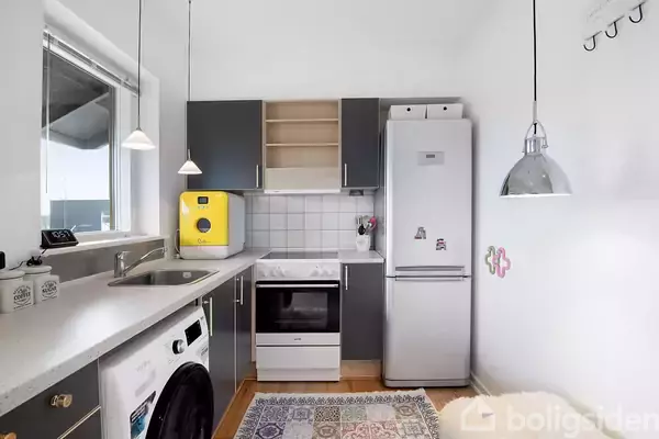 Køkken med grå skabe, køleskab, komfur og vaskemaskine. Gult mikroovn står på bordpladen. To hængelamper og en tæppe på gulvet. Vinduet giver lys til rummet.