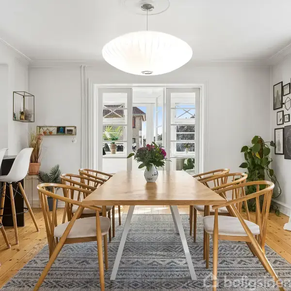 Et træbord med otte stole står midt i et lyst spiseområde. Over bordet hænger en stor, rund lampe. Rummet er omgivet af hvide vægge og grønne planter.