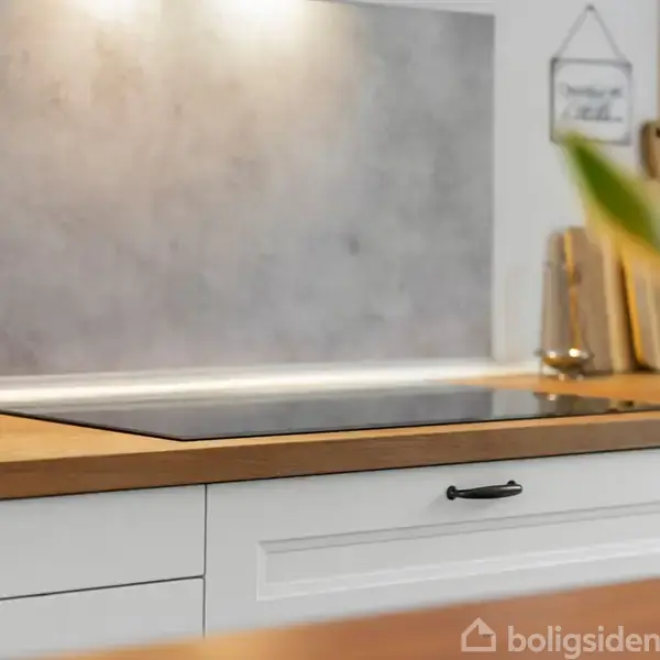 Kogeplade med træbordplade, flankeret af krydderikrukker og olier i træholder. I baggrunden ses skærebrætter og dæmpet belysning over grå vægbeklædning med to små rammer.