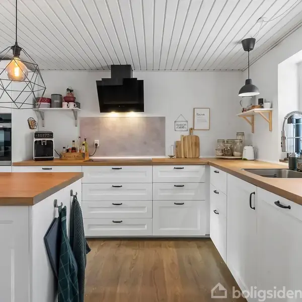 Et moderne køkken med hvidt interiør har træbordplader. Ovn og emhætte er indbygget. Lamper hænger fra loftet, og der er planter og køkkenudstyr. Panelet lyder: "This kitchen is for dancing."