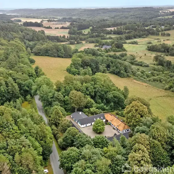 En stor ejendom omgivet af tæt skov ses fra oven. Ejendommen ligger i et frodigt, kuperet landskab med adskillige marker og skove i baggrunden.