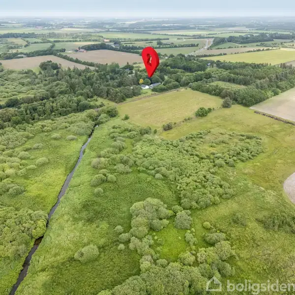 En rød kortnål markerer en position midt i et frodigt, landligt landskab. Der er skove, marker og en lille å, der skærer gennem området.