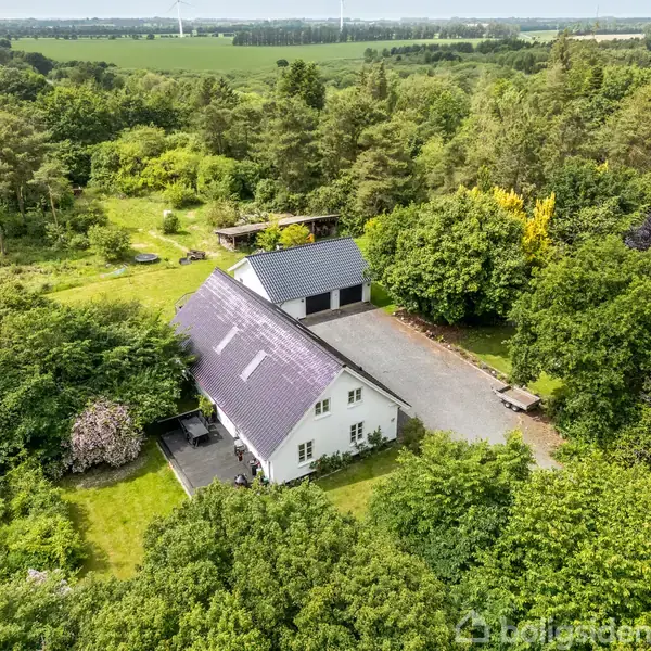 Et hvidt, landligt hus med mørkt tag ligger omgivet af frodig vegetation og skov. En separat garagebygning står tæt på. Grusvej og naturligt landskab præger baggrunden.