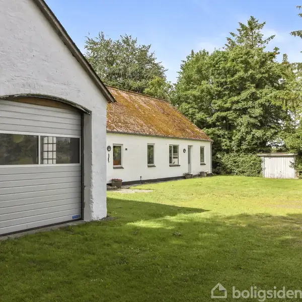 Huset er hvidt med et orange tag og har en garage med en port. Det står på en grøn græsplæne omgivet af træer og en blå himmel.