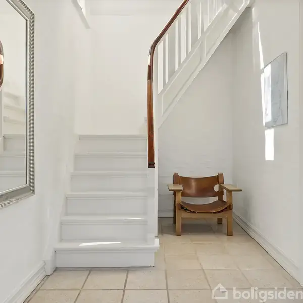 En stol står stille i en entré med hvide vægge og fliser. Til venstre er der en hvid trappe med et trægelænder og et stort spejl.