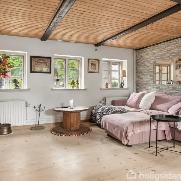 Stue med pink sofa ved vindue, sofabord foran, planter og billeder på væggen. Træloft med synlige bjælker. Stenvæg med spejlåbning til køkkenet i hjørnet.
