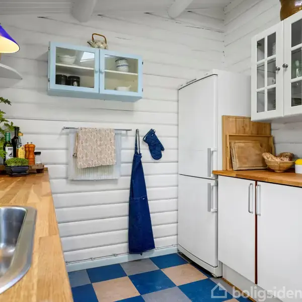 Køkken med træbordplade, vask og blå/hvid ternet gulv. Et køleskab står ved siden af et komfur. Ovenfor hænger et blåt vægskab og en lampe. Masser af køkkenudstyr og planter.