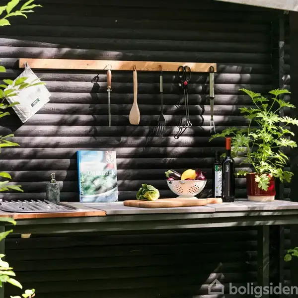Udendørs arbejdsbænk med træskeer, knive og redskaber hængende på en væg. På bordet er skærebrætter, en skål med frugt, en flaske olivenolie og en grøn plante.