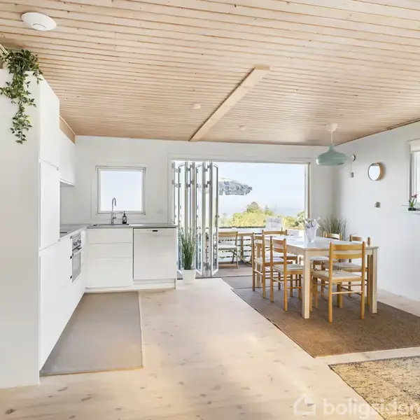 Et lyst køkken-alrum med hvidt køkken i venstre side, spisebord i træ til højre nær vinduer. Glidedøre åbner ud til balkon med udsigt til vand og grøn natur.