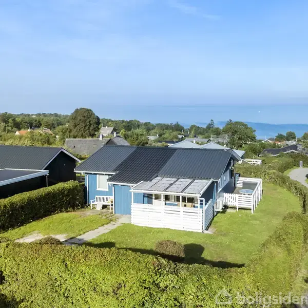 Blåt hus med terrasse står stille i en frodig have omgivet af hække; i baggrunden ses en snoet vej og havudsigt under klar blå himmel.