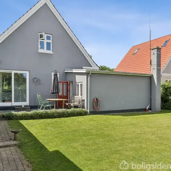 En gråmalet husfacade med et stejl tag har en lille terrasse med havemøbler og parasol. Husets anlagt baghave har en velplejet græsplæne og stenbelagt gangsti.