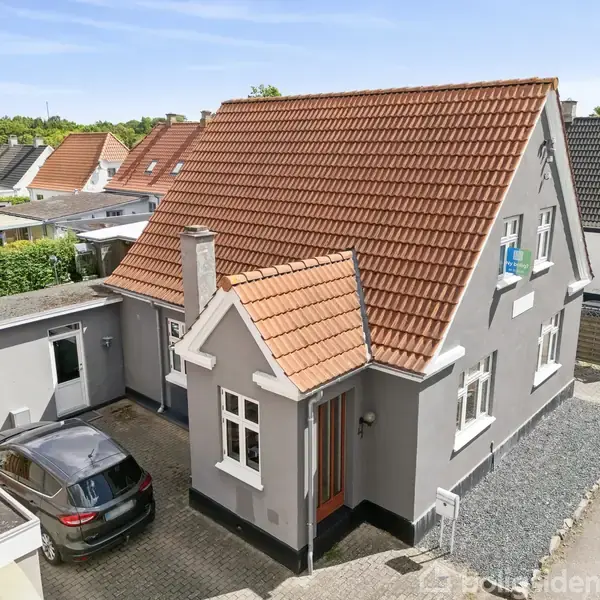 Et gråt hus med rødt tegltag står stille på en villavej, omgivet af andre huse. En sort bil er parkeret i indkørslen, og haven er begrønnet.