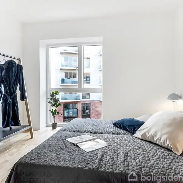 En seng med gråt sengetæppe har puder og en magasin på sig, belyst af en lille lampe. En stumtjener med tøj står i det lyse soveværelse ved vinduet.