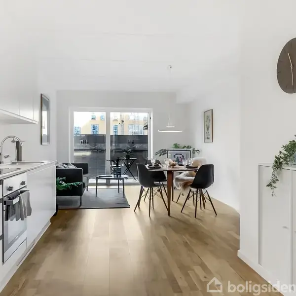 Køkken med hvidt interiør strækker sig mod en spiseplads med fire sorte stole omkring et bord. En sort sofa står ved vinduet i baggrunden. Vægur hænger til højre.