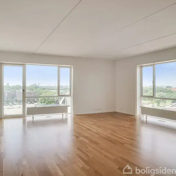 Stue med trægulv, store vinduer med udsigt til grønne omgivelser og en klar himmel. To radiatorer under vinduerne. Rummeligt og lyst med hvide vægge og loft.