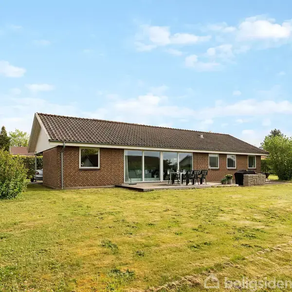 En murstensbungalow står stille i en velplejet have med græsplæne. En terrasse med havemøbler ses foran huset under en let skyet blå himmel.