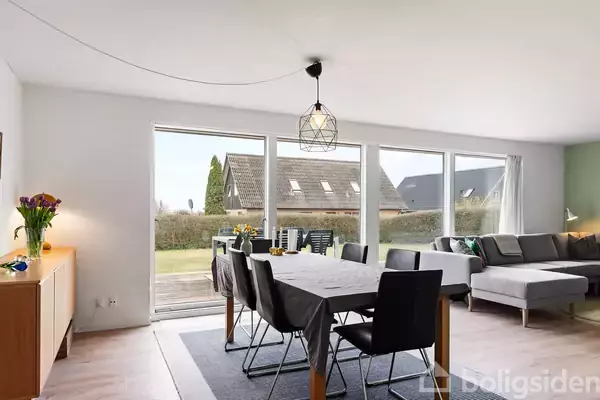 En spisebord med sorte stole står dækket i en stue. Store vinduer giver udsigt til en have og nabohuse med skrå tage. En sofa er placeret i hjørnet.