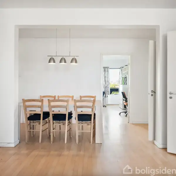 Seks træstole står ved et spisebord i et lyst rum med hvide vægge. Over bordet hænger tre loftslamper. Åben dør fører til et rum med vindue og skrivebord.