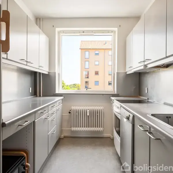 Et smalt køkken med hvide skabe, en vask og opvaskemaskine. Rummet har et vindue med udsigt til en anden bygning og grønne træer udenfor.