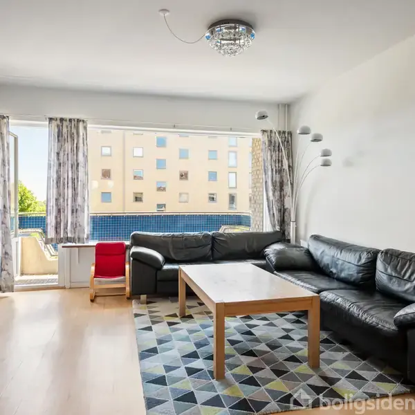 En stue med en stor, sort hjørnesofa står ved et sofabord. Rummet har et tæppe med geometrisk mønster, en lampe, et vindue med gardiner og udsigt til en bygning udenfor.