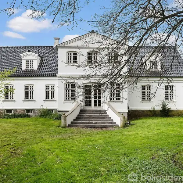 Hvid villa med sort tag står i centrum, omgivet af en grøn græsplæne med træer i forgrunden. Trapper fører til hovedindgangen. Himmel er klar med få skyer.
