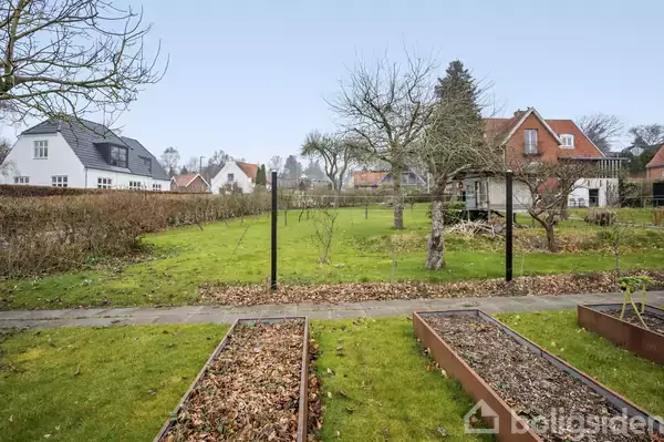 Hævede plantekasser står tomme i en grøn have. Nøgne træer og buske omgiver området. I baggrunden ses charmerende, hvide og røde huse med små haver.