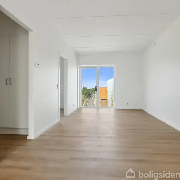 Et rummeligt, hvidmalet værelse med trægulv strækker sig mod en stor glasdør, som fører ud til en altan med udsigt over en grøn natur. I venstre side ses en indbygget skab.